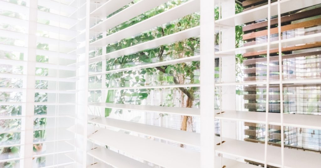 White Shutters Partially Open, Revealing A View Of Green Foliage Outdoors