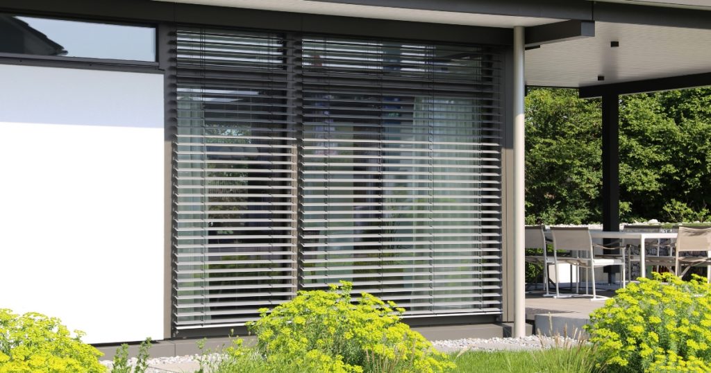 Modern House With Large Windows Covered By Dark Gray Horizontal Blinds, Surrounded By A Garden With Yellow Flowers